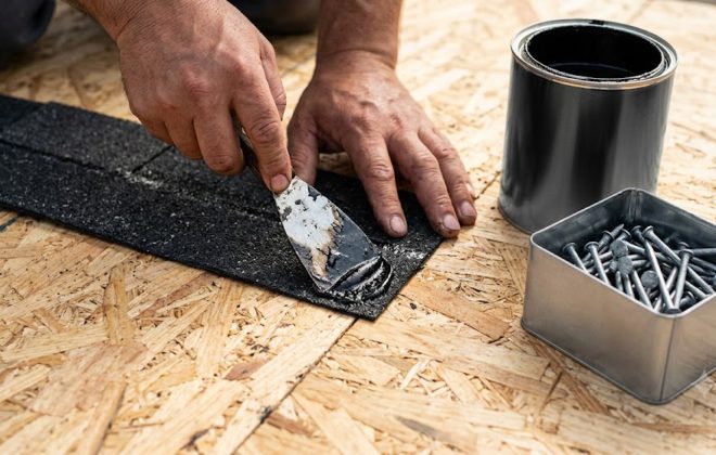 Instalador aplicando adhesivo asfaltico con espatula sobre palmeta de teja asfaltica junto a clavos de cabeza ancha en tablero OSB